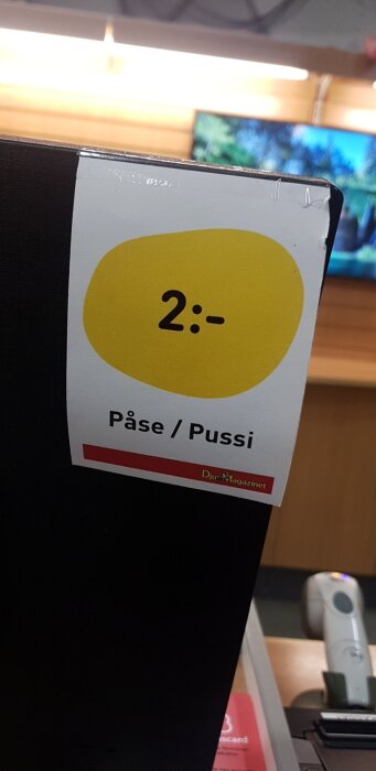 Etikett på svart yta visar pris 2 kronor för "Påse/Pussi" i en butiksmiljö med datorskärm i bakgrunden.