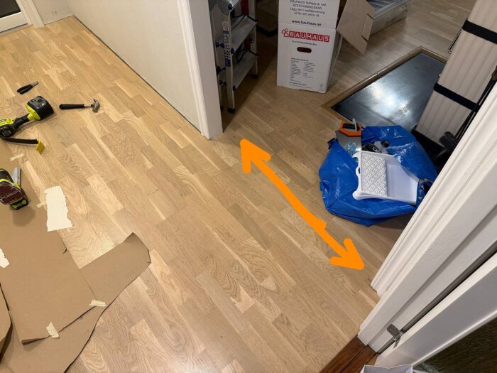 Verktyg och material på ett laminatgolv mellan hall och kök; en blå IKEA-påse med verktyg syns i bakgrunden och en orange pil markerar övergången.