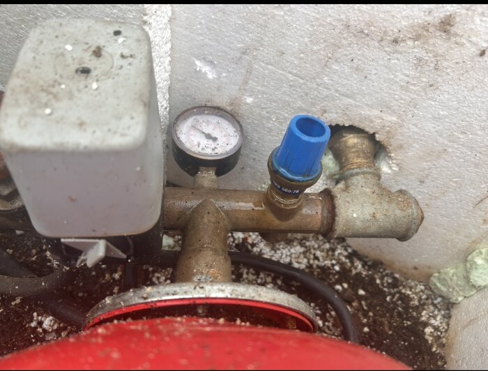 VVS-komponenter med en blå ventil på ett rör, en tryckmätare och en tryckströmbrytare, omgivna av murverk och smuts.