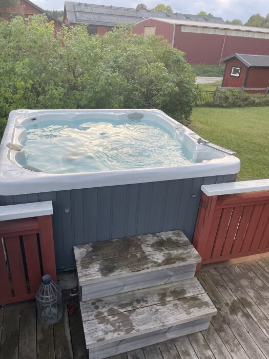 Utomhusjacuzzi omgiven av grönska med trätrappa och röd byggnad i bakgrunden.