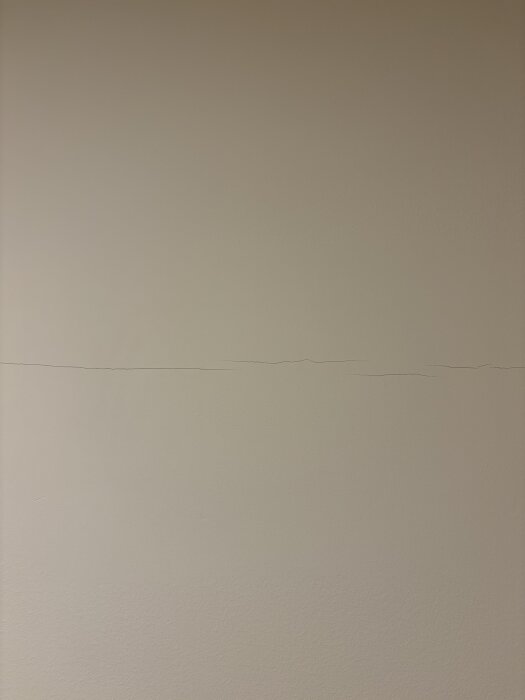 Spricka i ljus innervägg, cirka 1 mm bred, lodrätt, synlig på gipsskivans skarv.