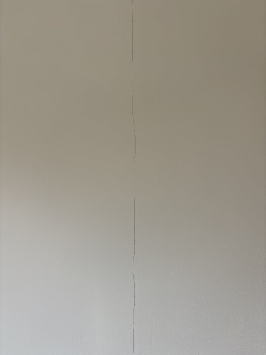 Spricka på ljus innervägg, cirka 1 mm bred, vertikalt längs gipsskarv.