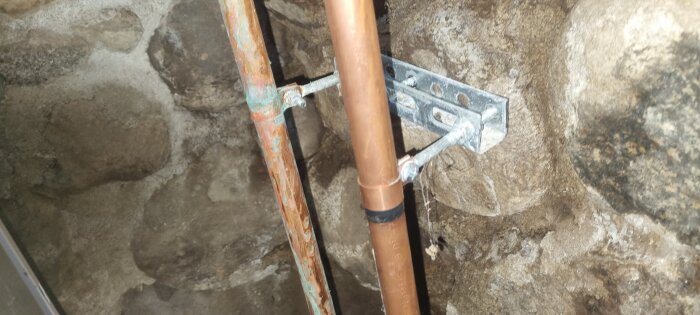 Två kopparrör monterade på en stenvägg, det vänstra rören har korrosion och det högra verkar nytt med eltejp samt metallklammer som fäster dem.