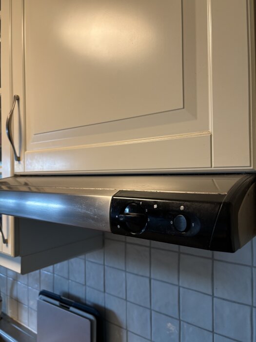 Köksfläkt i rostfritt stål under skåp, med svart kontrollpanel och vred, i ett radhuskök med kaklad vägg i bakgrunden.