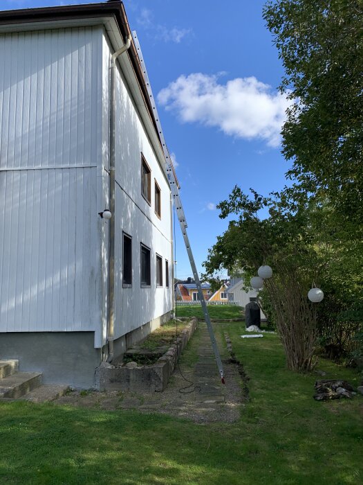 Stege lutad mot tvåvåningshus under blå himmel, träd vid sidan och trädgård nedanför.