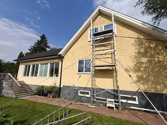 Ställning uppsatt vid gul husfasad för målning av vindskivor och byte av gavelsprång. Stödd med spännband för stabilitet.