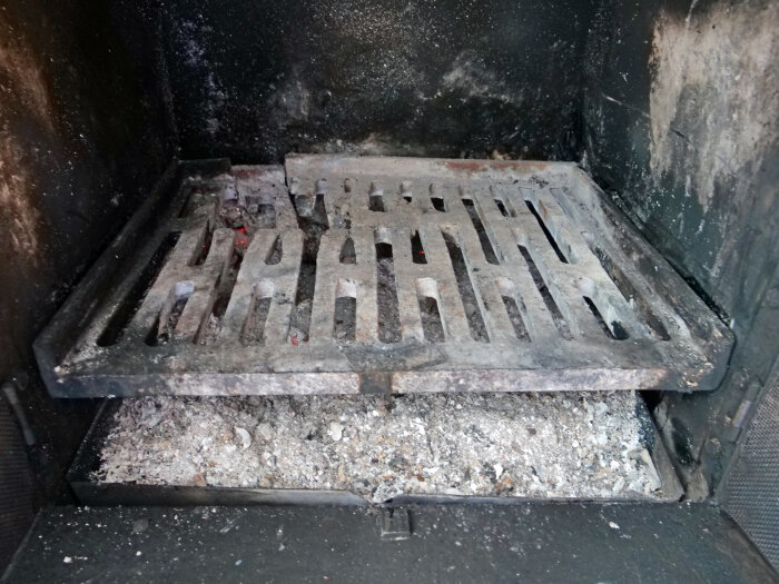 Rostigt och delvis trasigt metallgaller i en eldstad, omgivet av sot och aska. Gallerplattan har sprickor och visar tecken på slitage.