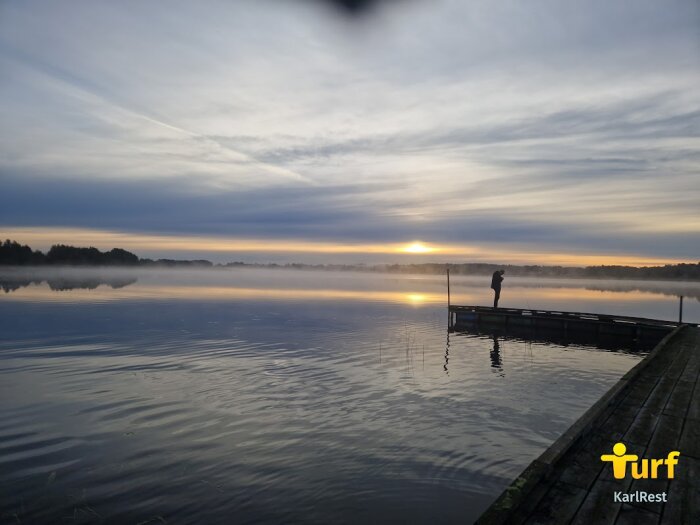 Solnedgång över en sjö med en person på brygga vid Hammarö, dimma över vattnet reflekterar de gyllene solstrålarna.