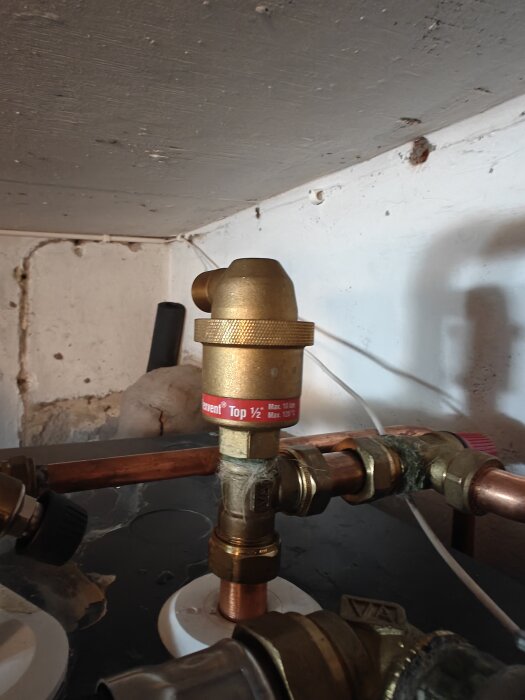 Ventil ansluten till kopparrör med rörkopplingar, placerad i en källare med synlig tak- och väggstruktur i bakgrunden.