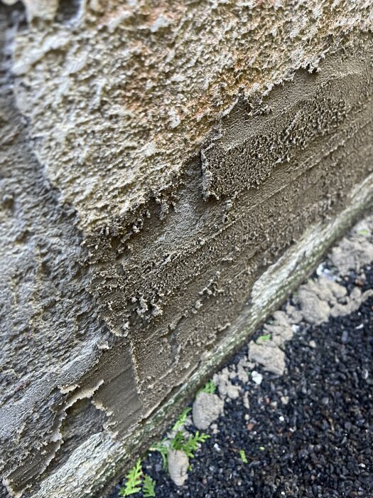 Putsad husgrund med ojämn yta där bruket inte fäster ordentligt. Grus och små växter syns vid basen.