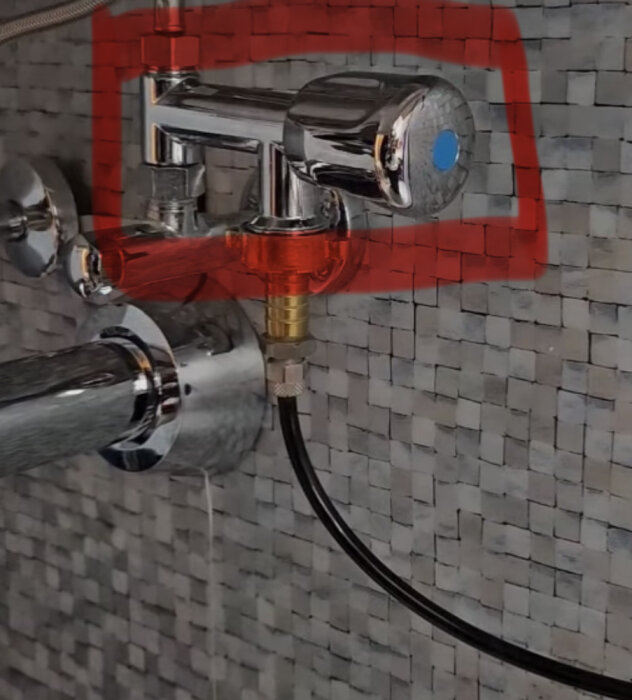 Kromad T-koppling med ventil mot mosaikvägg i badrum, ansluten till svart slang och annan rörledning. Röd markering runt ventilen.