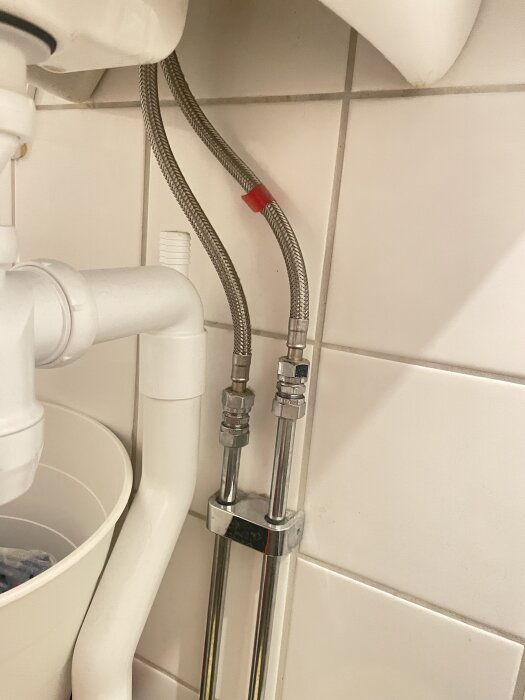 Två metallslangar och rör under badrumshandfat anslutna med ventiler, mot en kaklad vägg.