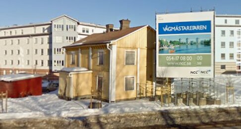 Gult tvåvåningshus före renovering, med en stor skylt intill som annonserar "Vägmästarearenan." Snö på marken och omgivande byggnader i bakgrunden.