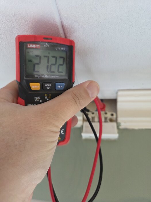 Multimeter visar 27,22 volt, hålls av en hand nära ett vägguttag.