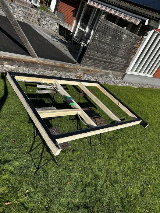 Träram för sänggavel under konstruktion utomhus, placerad på gräsmatta, omgiven av trädgårdsmöbler och husfasad i bakgrunden.