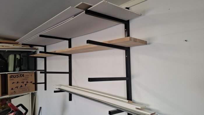 Vägghängd virkesförvaring i svart metall och trä, svetsad och monterad i ett garage. Fylld med träbitar och byggmaterial.