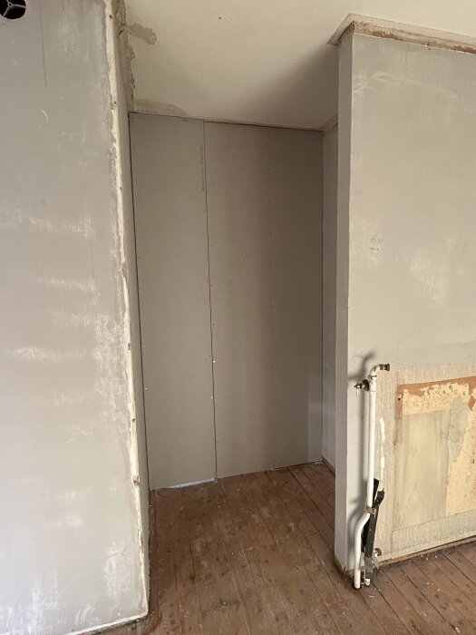 Inbyggd dörröppning i hall med grå väggar och trägolv, tidigare ingång till kök. Synliga rör på högra väggen.