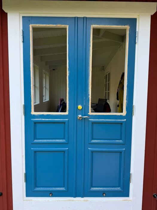 Blå dubbeldörr med borttagna ytterfönster, omgiven av vit och röd panel. Förberedelser för fräsning och slipning syns i karmarna.