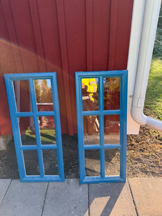 Blåmålade ytterfönster lutar mot en röd vägg, bredvid stuprör och trädgård.