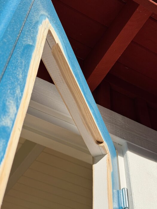 Fräst och slipat ytterfönster med blå tejp, placerat öppet under ett rött tak. Synlig trästruktur och metallgångjärn.