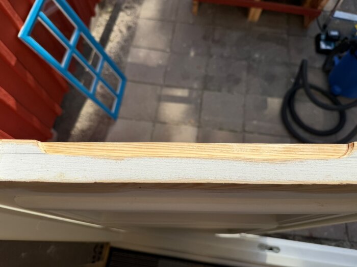 Ytterfönster med fräsning längs kanten, liggande på sidan, blå fönsterkarm i bakgrunden mot röd vägg, soligt väder.