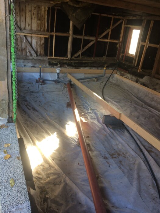 Byggarbetsplats med plastmatta, avloppsrör och träreglar, del av renoveringsprojekt.