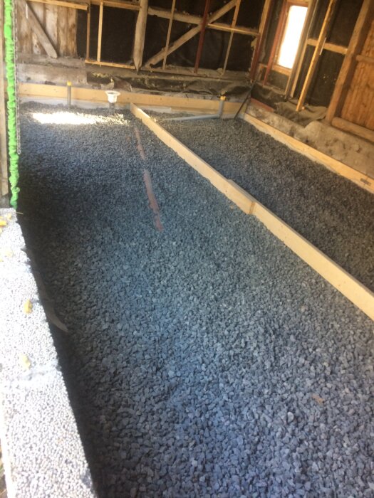 Byggarbetsplats med en grund av grus och träformar i ett oskyddat rum med exponerade träväggar och fönster.