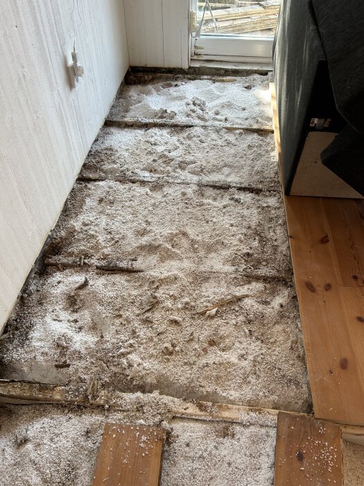 Golvet med upplöft trägolv och sågspån, synlig isolering och vägguttag, vid ett rum med dörr mot terrassen.