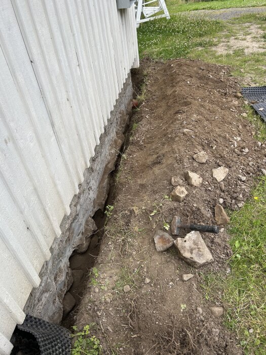 Djupt grävt dike längs en byggnadsgrund med verktyg och stenblock vid sidan av.