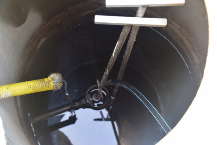 Brunn med två spakar och rör, vatten synligt i botten, gul kabel och ventiler observerade under inspektionslucka.