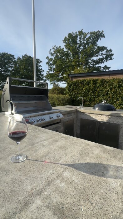 Utekök med grill under klar himmel och ett glas rött vin på en bänk.