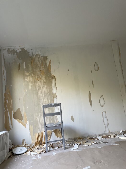 En vägg med avskavd tapet, en stol framför och rivna tapetbitar på golvet.