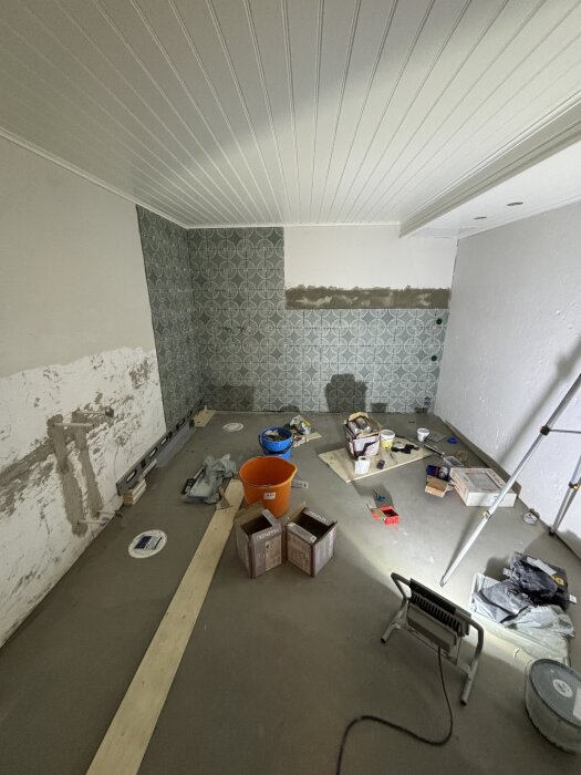 Renovering av rum med mönstrade väggar. Byggmaterial och verktyg spridda över golvet; väggen är nästan klar.
