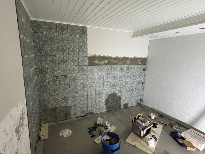 Renovering pågår: kaklad vägg med mönster och oordning av byggmaterial på golvet i ett rum under konstruktion.