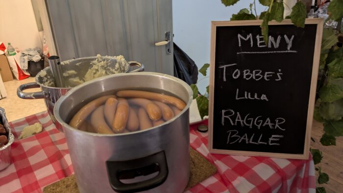 Gryta med kokta korvar på rutig duk bredvid griffeltavla med texten "MENY Tobbe's Lilla Raggar Balle".