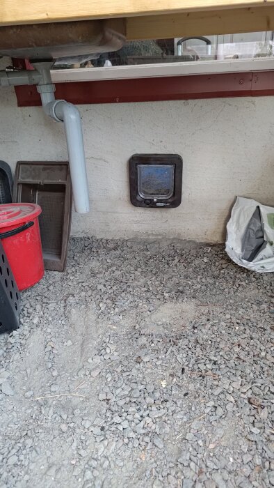 Kattlucka installerad i en vägg med grus på marken. Plastkärl och rör syns bredvid.