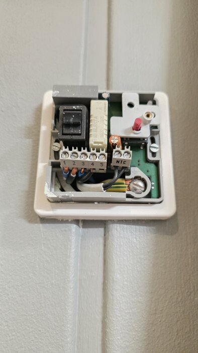 Öppen golvvärmetermostat monterad på väggen, med synliga kablar och komponenter, saknar märkning och modellinformation.