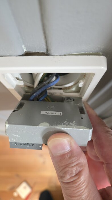 En person håller i en termostat för golvvärme, delvis utdragen från väggen med synliga kablar bakom.