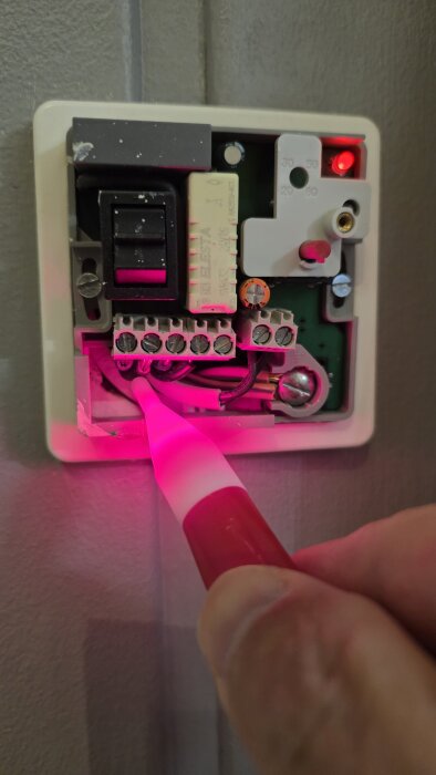 En elektrisk termostat med exponerade ledningar undersöks med en testpenna. En röd lampa lyser på termostatens framsida.