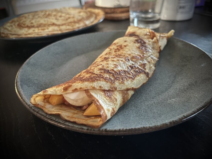 Pannkaka fylld med fruktsallad (persika, plommon, jordgubbar, mango, satsumas, äpple) och en klick grädde på en grå tallrik.