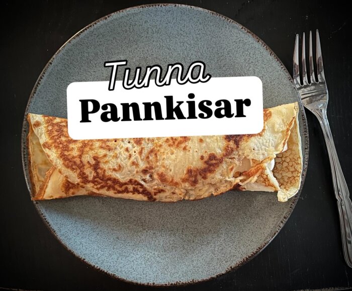 Tunn pannkaka serverad på en tallrik, med texten "Tunna Pannkisar" i bildens mitt, och en gaffel placerad bredvid tallriken.