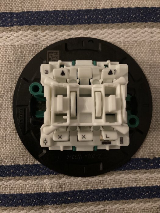 Elektrisk väggmonterad brytare av plast med vit och svart komponenter placerad på en randig tygbakgrund.