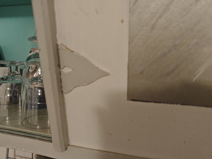 Ett närbildsfoto av en vit skåplucka med avskavt hörn och glas på en hylla i bakgrunden.