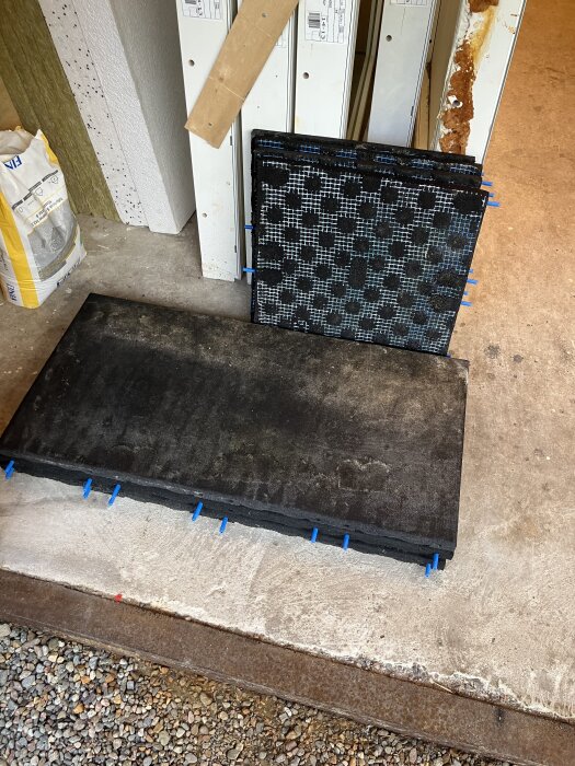 Svarta gymmattor staplade i garage, några med mönster. Ligger på betonggolv bredvid byggmaterial och säck.