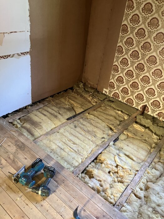 Rivet sovrumsgolv med synlig isolering, gamla tapeter på väggarna, och två borrmaskiner på trägolvet i förgrunden.