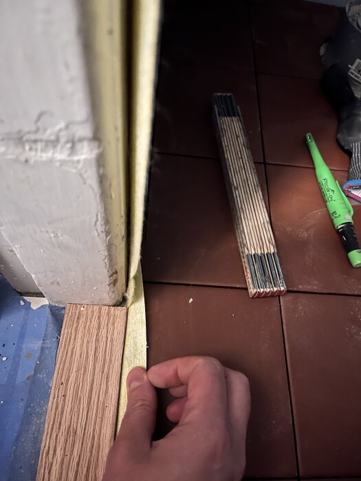 Hand håller i tejp vid dörrtröskel av ek mot ljusa dörrfoder och bruna klinkerplattor.