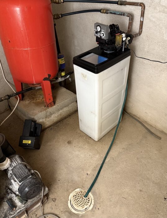 Vit plastbehållare bredvid en röd trycktank i ett pumprum, med en slang som leder till ett golvavlopp och olika röranslutningar.