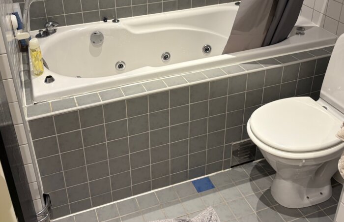 Badkar inklätt med grått kakel, duschmunstycke och toalett till höger, ingen synlig servicelucka.