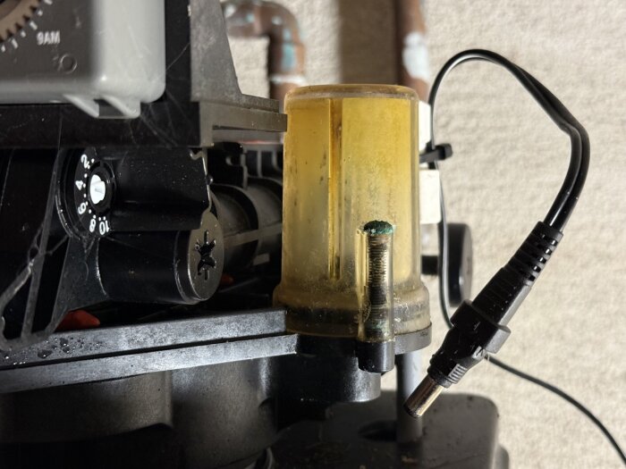 Gult och smutsigt filter kopplat till en vattensystemanordning, med anslutna kablar och ett vred, delvis täckt av smuts och korrosion.