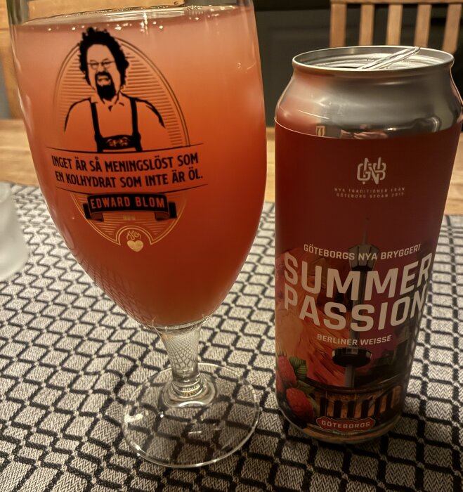 Ett glas med Summer Passion öl bredvid en ölburk på ett mönstrat underlägg. Glaset har ett tryck med Edward Blom och text om kolhydrater.